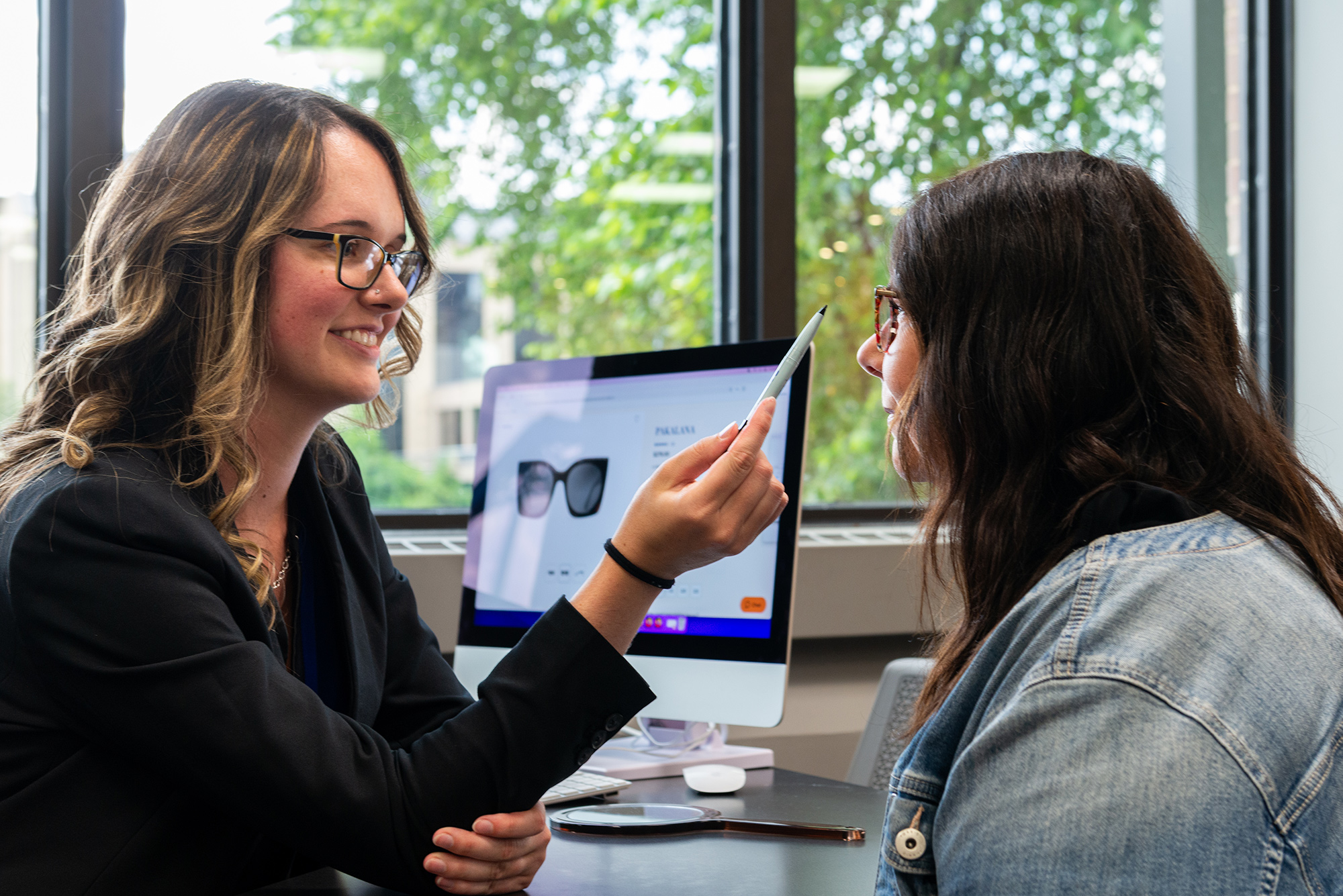 An optometrist helps a patient try on glasses while sitting at a desk with a computer displaying eyewear options - Eye Doctor in Grand Rapids MI